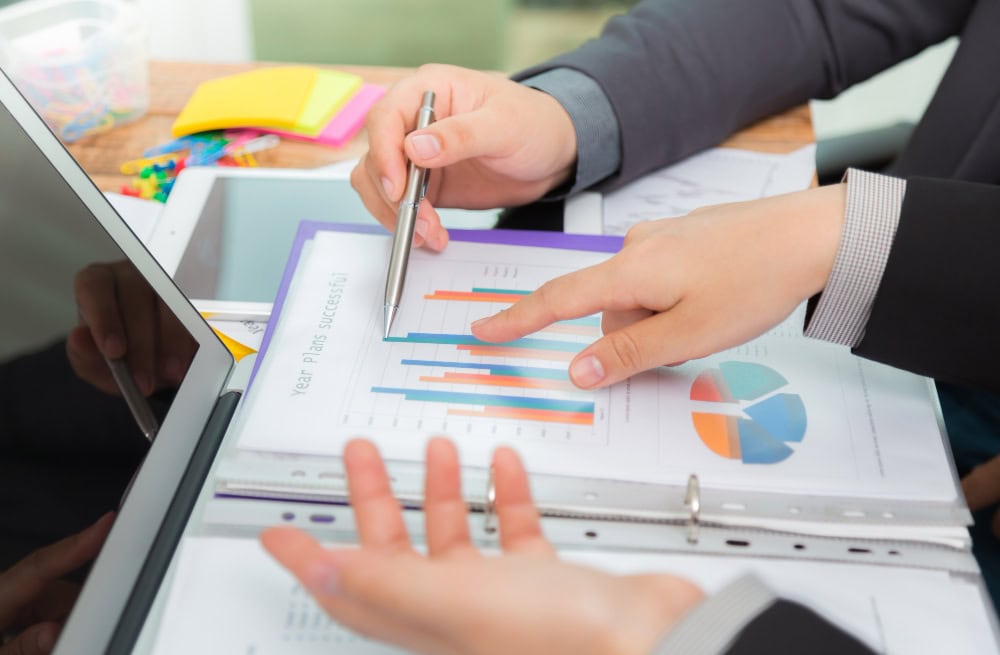 Two professionals review colorful charts and graphs in a binder during a meeting, with a laptop nearby.