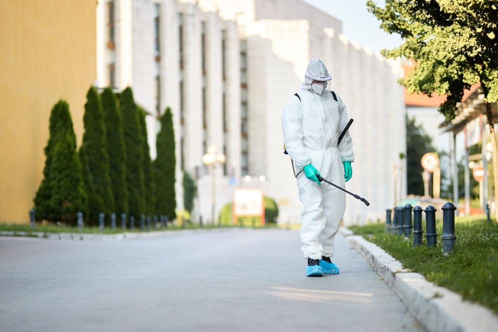 Person in protective suit and mask spraying disinfectant on city street, showing fast emergency pest control response.