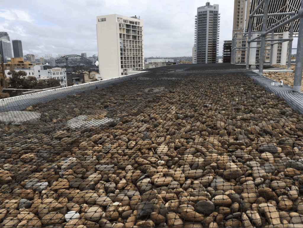 Flat rooftop with pebbles and black netting for high rise bird proofing, tall buildings behind, cloudy sky overhead.