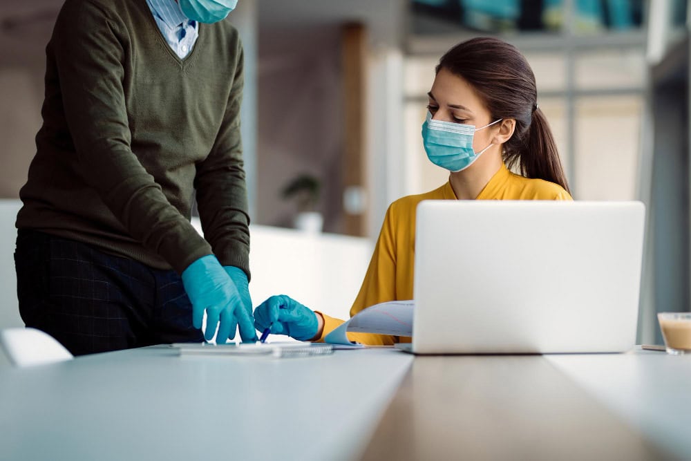 Two people in masks and gloves discuss preventative rodent pest control at an office desk with a laptop and documents.