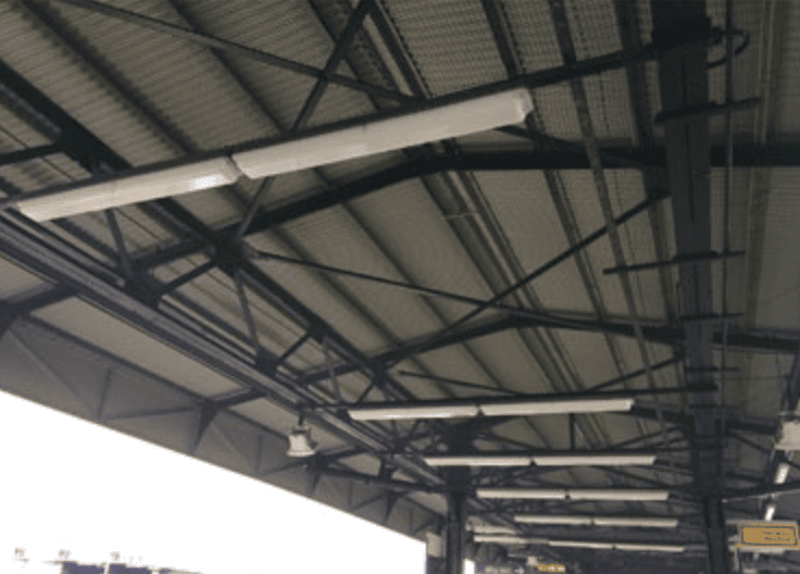 Industrial ceiling with dark steel beams, pipes, and suspended fluorescent lights in a warehouse or station setting.