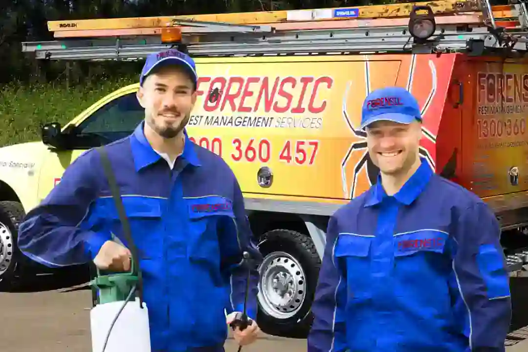Two pest control workers in blue uniforms stand by a yellow Forensic Pest Management van, smiling with equipment.